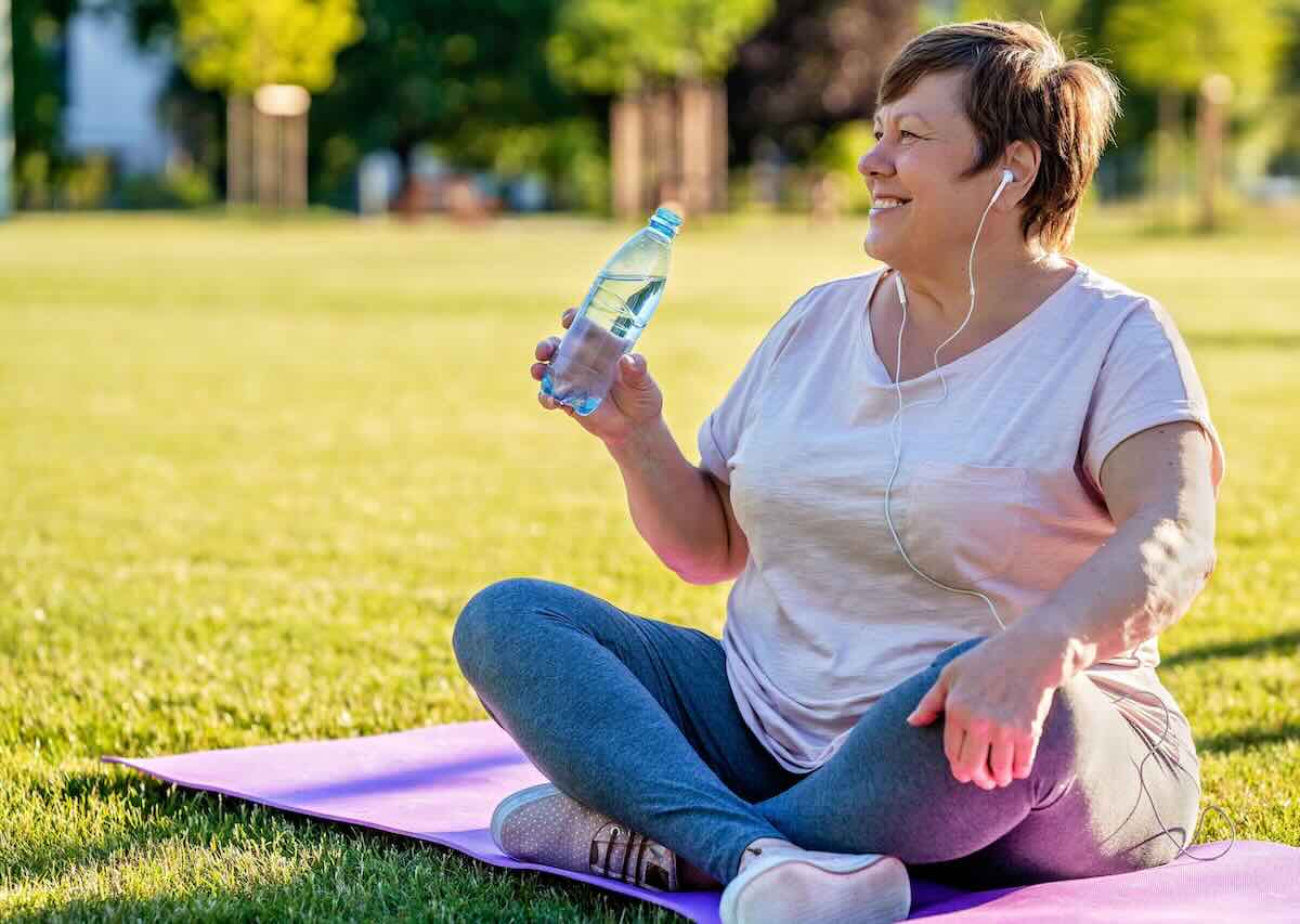 Woman relaxing on a yoga mat in the park, holding a water bottle after exercise.